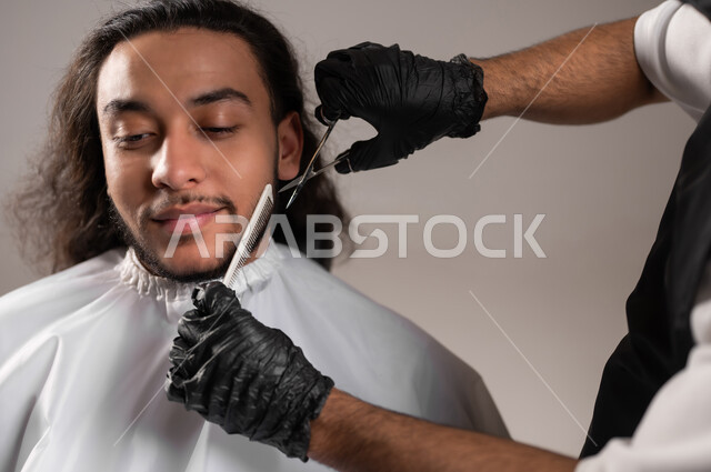 ممارسة مهنة الحلاقة, تقديم خدمات العناية الشخصية, الاهتمام بالشعر و النظافة البدنية, بروتريه مقرب لحلاق عربي خليجي يرتدي زي الحلاقين التقليدي مع القفازات يحلق لشاب سعودي, استخدام الغطاء الأبيض لحماية الملابس, خلفية رمادية