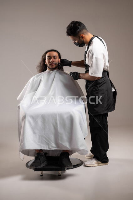 ممارسة مهنة الحلاقة, تقديم خدمات العناية الشخصية, الاهتمام بالشعر و النظافة البدنية, بروتريه لحلاق عربي خليجي يرتدي زي الحلاقين التقليدي مع الكمامة يحلق لشاب سعودي, استخدام الغطاء الأبيض لحماية الملابس, الجسم بالطول الكامل, خلفية رمادية