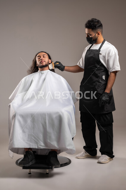 استخدام الغطاء الأبيض لحماية الملابس, الاهتمام بالشعر و النظافة البدنية, تقديم خدمات العناية الشخصية, بورتريه لحلاق عربي خليجي يرتدي زي الحلاقين التقليدي مع الكمامة يحلق لشاب سعودي, الجسم بالطول الكامل, خلفية رمادية