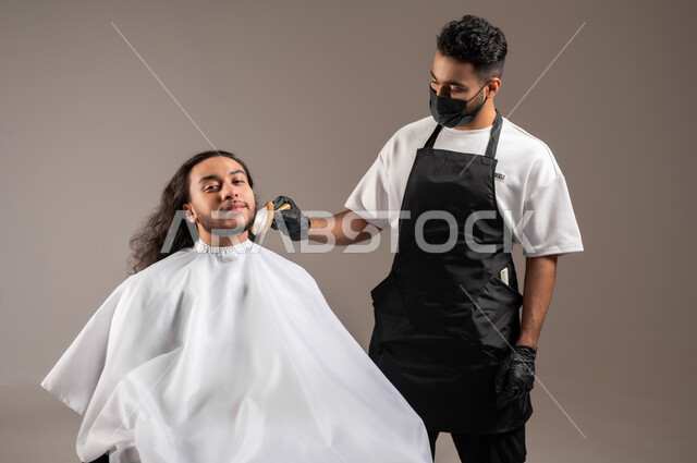 استخدام الغطاء الأبيض لحماية الملابس, الاهتمام بالشعر و النظافة البدنية, تقديم خدمات العناية الشخصية, بورتريه لحلاق عربي خليجي يرتدي زي الحلاقين التقليدي مع الكمامة يحلق لشاب سعودي, خلفية رمادية