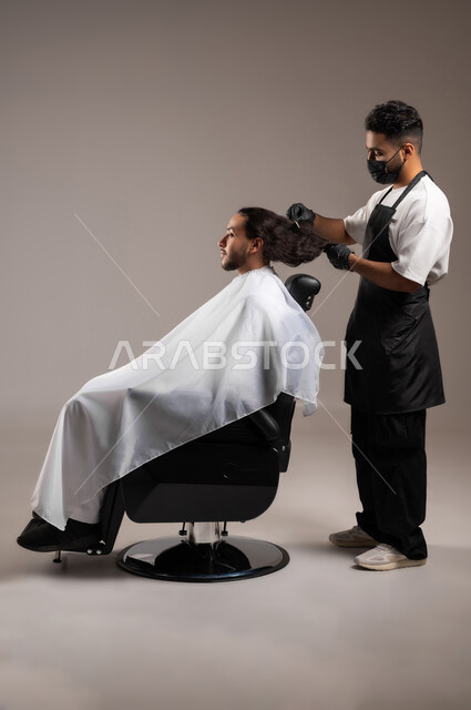 تقديم خدمات العناية الشخصية, الاهتمام بالمظهر الخارجي و النظافة البدنية, بروتريه لحلاق عربي خليجي يرتدي زي الحلاقين لتقليدي مع الكمامة يقوم بتمشيط شعر شاب سعودي, استخدام الغطاء الأبيض لحماية الملابس, الجسم بالطول الكامل, خلفية رمادية