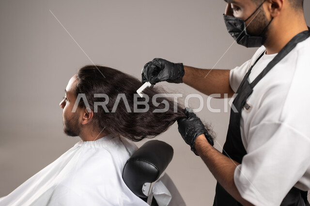 تقديم خدمات العناية الشخصية, الاهتمام بالمظهر الخارجي و النظافة البدنية, بروتريه مقرب لحلاق عربي خليجي يرتدي زي الحلاقين لتقليدي مع الكمامة يقوم بتمشيط شعر شاب سعودي, استخدام الغطاء الأبيض لحماية الملابس, خلفية رمادية