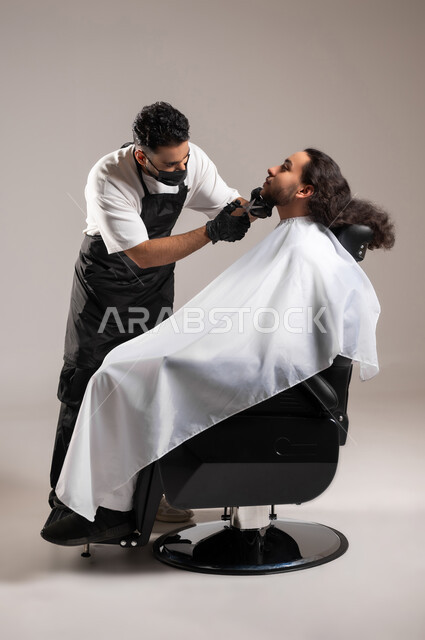 تقديم خدمات العناية الشخصية, استعمال المقص و المشط لحلاقة الذقن, بورتريه لحلاق عربي خليجي يرتدي زي الحلاقين التقليدي مع الكمامة يحلق لشاب سعودي, استخدام الغطاء الأبيض لحماية الملابس, الجسم بالطول الكامل, خلفية رمادية