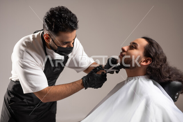 Providing personal care services, using scissors and a comb to shave the beard, a close-up portrait of a Gulf Arab barber wearing a traditional barber uniform with a mask, shaving a Saudi young man, using a white cover to protect clothes, gray background