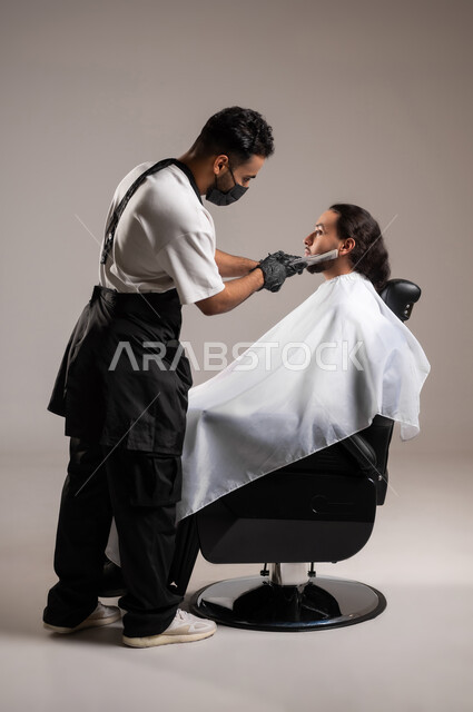 استعمال المقص و المشط لحلاقة الذقن, تقديم خدمات العناية الشخصية, بورتريه لحلاق عربي خليجي يرتدي زي الحلاقين التقليدي مع الكمامة يحلق لشاب سعودي, استخدام الغطاء الأبيض لحماية الملابس, الجسم بالطول الكامل, خلفية رمادية