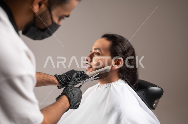 استعمال المقص و المشط لحلاقة الذقن, تقديم خدمات العناية الشخصية, بورتريه مقرب لحلاق عربي خليجي يرتدي زي الحلاقين التقليدي مع الكمامة يحلق لشاب سعودي, استخدام الغطاء الأبيض لحماية الملابس, خلفية رمادية