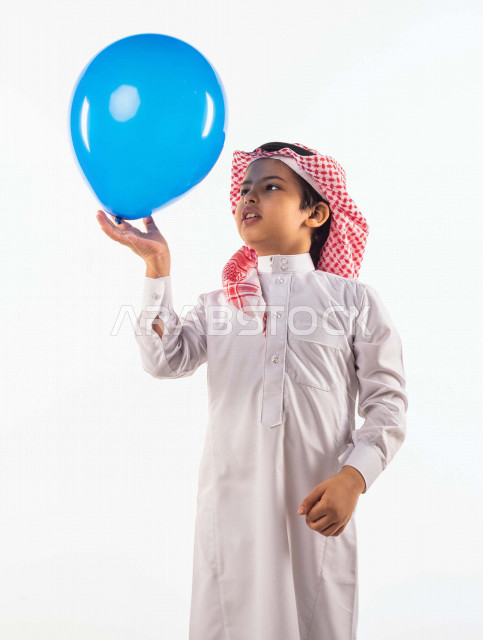 خلفية بيضاء لصبي صغير سعودي خليجي مبتسم وجميل ، يلعب بالبالون أزرق اللون بإيماءات وجه ويدين مختلفة تدل على السعادة , بورتريه لطفل سعودي خليجي سعيد يرتدي الزي السعودي التقليدي , الاحتفال بعيد الفطر السعيد ،عيد الفطر المبارك ,التسليه والترفيه