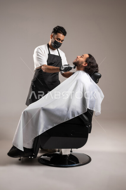 تقديم خدمات العناية الشخصية, الاهتمام بالشعر و النظافة البدنية, استعمال ماكينة الحلاقة, بورتريه لحلاق عربي خليجي يرتدي زي الحلاقين التقليدي مع الكمامة يحلق لشاب سعودي, استخدام الغطاء الأبيض لحماية الملابس, الجسم بالطول الكامل, خلفية رمادية