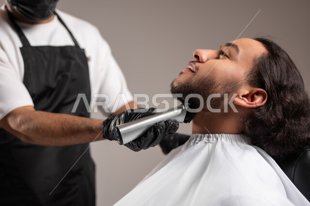تقديم خدمات العناية الشخصية, الاهتمام بالشعر و النظافة البدنية, استعمال ماكينة الحلاقة, بورتريه مقرب لحلاق عربي خليجي يرتدي زي الحلاقين التقليدي مع الكمامة يحلق لشاب سعودي, استخدام الغطاء الأبيض لحماية الملابس, خلفية رمادية