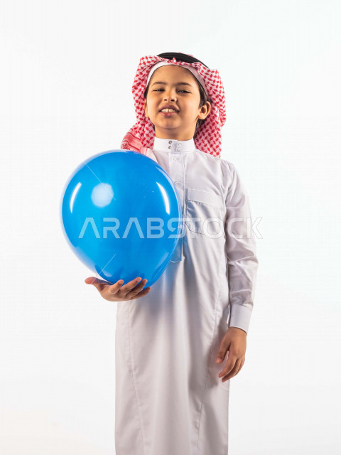 خلفية بيضاء لصبي صغير سعودي خليجي مبتسم وجميل ، يلعب بالبالون أزرق اللون بإيماءات وجه ويدين مختلفة تدل على السعادة , بورتريه لطفل سعودي خليجي سعيد يرتدي الزي السعودي التقليدي , الاحتفال بعيد الفطر السعيد ،عيد الفطر المبارك ,التسليه والترفيه