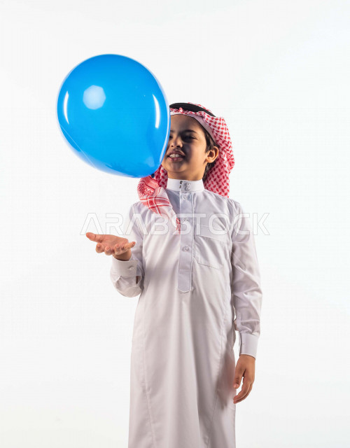 خلفية بيضاء لصبي صغير سعودي خليجي مبتسم وجميل ، يلعب بالبالون أزرق اللون بإيماءات وجه ويدين مختلفة تدل على السعادة , بورتريه لطفل سعودي خليجي سعيد يرتدي الزي السعودي التقليدي , الاحتفال بعيد الفطر السعيد ،عيد الفطر المبارك ,التسليه والترفيه