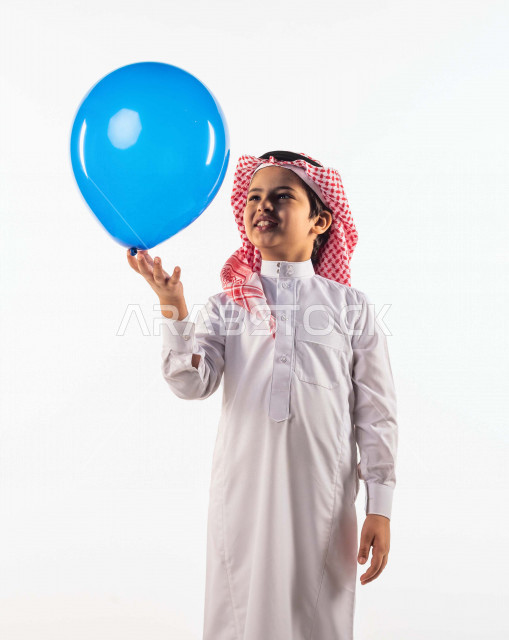 خلفية بيضاء لصبي صغير سعودي خليجي مبتسم وجميل ، يلعب بالبالون أزرق اللون بإيماءات وجه ويدين مختلفة تدل على السعادة , بورتريه لطفل سعودي خليجي سعيد يرتدي الزي السعودي التقليدي , الاحتفال بعيد الفطر السعيد ،عيد الفطر المبارك ,التسليه والترفيه
