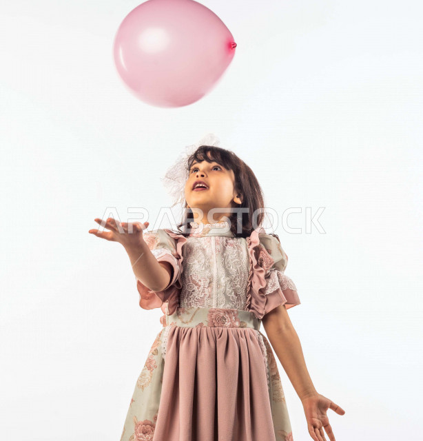 خلفية بيضاء لفتاه صغيرة سعودية خليجية مبتسمة وجميلة ، تلعب بالبالون زهري اللون بإيماءات وجه ويدين مختلفة تدل على السعادة , بورتريه لطفلة سعودية خليجية سعيدة ترتدي لباس العيد الانيق , الاحتفال بعيد الفطر السعيد ، عيد الفطر المبارك , التسليه والترفيه