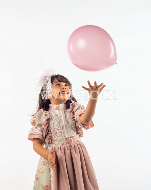 خلفية بيضاء لفتاه صغيرة سعودية خليجية مبتسمة وجميلة ، تلعب بالبالون زهري اللون بإيماءات وجه ويدين مختلفة تدل على السعادة , بورتريه لطفلة سعودية خليجية سعيدة ترتدي لباس العيد الانيق , الاحتفال بعيد الفطر السعيد ، عيد الفطر المبارك , التسليه والترفيه