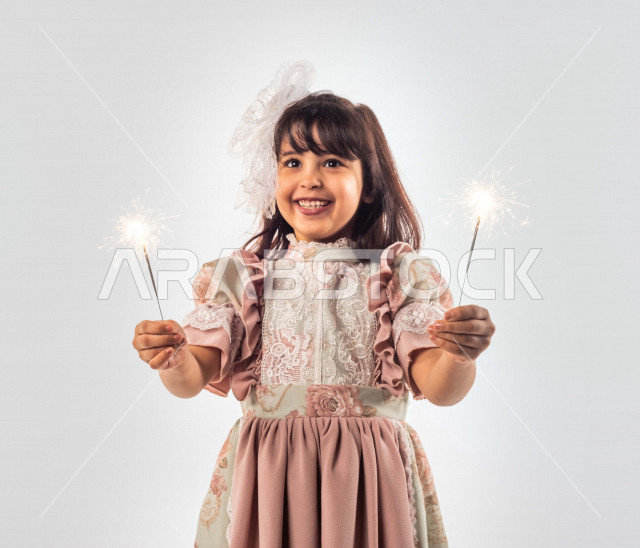 خلفية بيضاء لفتاه صغيرة سعودية خليجية مبتسمة وجميلة ، تمسك بيدها عصا الألعاب النارية السحرية بإيماءات وجه ويدين مختلفة تدل على السعادة , بورتريه لطفلة سعودية خليجية سعيدة ترتدي لباس العيد الانيق , الاحتفال بعيد الفطر السعيد ، عصا الألعاب النارية السحرية