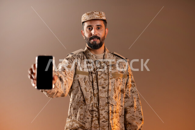 قوات التدخل السريع، الوقوف باستقامة واستعداد وانضباط، بورتريه لرجل عربي خليجي سعودي يرتدي الزي العسكري يشير بهاتفهه المحمول بشاشه سوداء فارغه، الحفاظ على الأمن والأمان، القوات المسلحه السعوديه، خلفيه برتقاليه