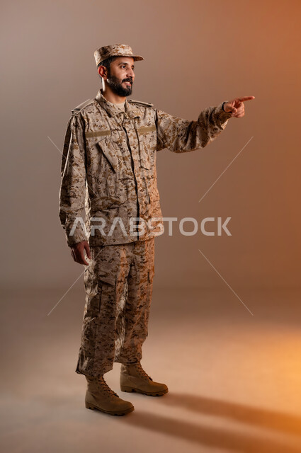 Movements indicating going somewhere, contemplating and setting goals accurately, portrait of a Saudi Gulf Arab man wearing a military uniform pointing with his hand to a specific place, standing and looking at a specific location and pointing with his index finger to reach the goal, Saudi Armed Forces, orange background