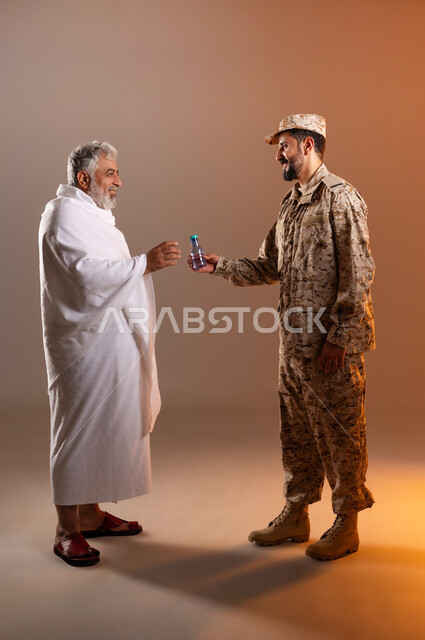 مساعده الحجاج والمعتمرين القادمين الى مكه المكرمه لاداء المشاعر الدينيه، شهامه رجال الامن في خدمه ضيوف الرحمن، بورتريه لرجل عربي خليجي سعودي يرتدي الزي العسكري يقوم بتقديم الماء لرجل كبير بالسن يرتدي ثياب الاحرام، خلفيه برتقاليه
