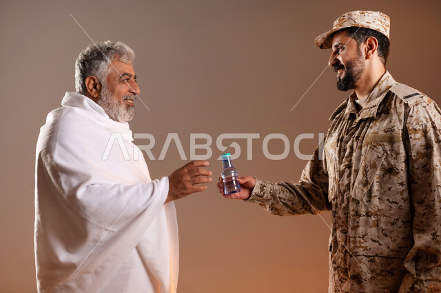 مساعده الحجاج والمعتمرين القادمين الى مكه المكرمه لاداء المشاعر الدينيه، شهامه رجال الامن في خدمه ضيوف الرحمن، بورتريه لرجل عربي خليجي سعودي يرتدي الزي العسكري يقوم بتقديم الماء لرجل كبير بالسن يرتدي ثياب الاحرام، خلفيه برتقاليه