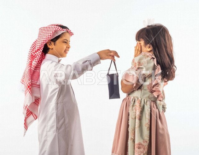 خلفية بيضاء لصبي وفتاه صغيران سعوديان مبتسمان ، الفتاه الصغيرة تضع يدها على خدها تشعر بالخجل , يمسك الطفل السعودي هدية بكيس أسود بإيماءات وجه ويدين مختلفة , بورتريه لطفلين سعوديان خليجيان سعيدان يرتديان لباس العيد الأنيق ,الاحتفال بعيد الفطر السعيد