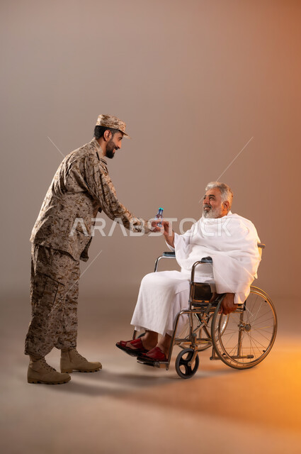مساعده الحجاج والمعتمرين القادمين الى مكه المكرمه لاداء المشاعر الدينيه، شهامه رجال الامن في خدمه ضيوف الرحمن، بورتريه لرجل عربي خليجي سعودي يرتدي الزي العسكري يقوم بتقديم الماء لرجل كبير بالسن يرتدي ثياب الاحرام مقعد على كرسي من ذوي الاحتياجات الخاصه، خلفيه برتقاليه
