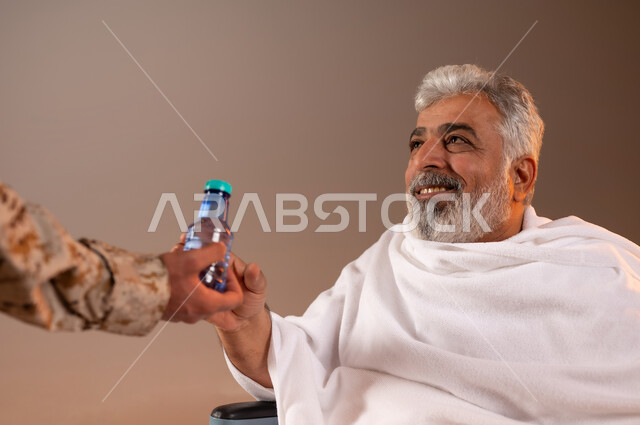 مساعده الحجاج والمعتمرين القادمين الى مكه المكرمه لاداء المشاعر الدينيه، شهامه رجال الامن في خدمه ضيوف الرحمن، بورتريه لرجل عربي خليجي سعودي يرتدي الزي العسكري يقوم بتقديم الماء لرجل كبير بالسن يرتدي ثياب الاحرام مقعد على كرسي من ذوي الاحتياجات الخاصه، خلفيه برتقاليه