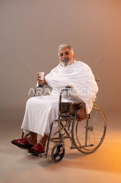 الاستعداد لاداء مناسك الحج والعمره، التقرب الى الله بالاعمال الصالحه، بورتريه لرجل عربي خليجي سعودي كبير بالسن يرتدي ثوب الاحرام يجلس على كرسي متحرك من ذوي الاحتياجات الخاصه يقوم بشرب ماء زمزم، الركن الخامس من أركان الاسلام لمن استطاع اليه سبيلا، خلفيه برتقاليه