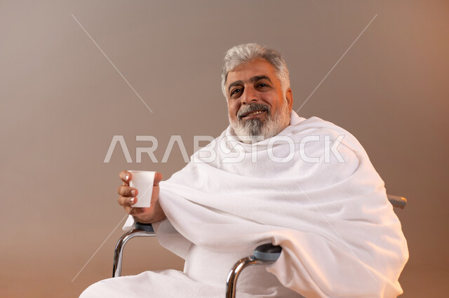 الاستعداد لاداء مناسك الحج والعمره، التقرب الى الله بالاعمال الصالحه، بورتريه لرجل عربي خليجي سعودي كبير بالسن يرتدي ثوب الاحرام يجلس على كرسي متحرك من ذوي الاحتياجات الخاصه يقوم بشرب ماء زمزم، الركن الخامس من أركان الاسلام لمن استطاع اليه سبيلا، خلفيه برتقاليه