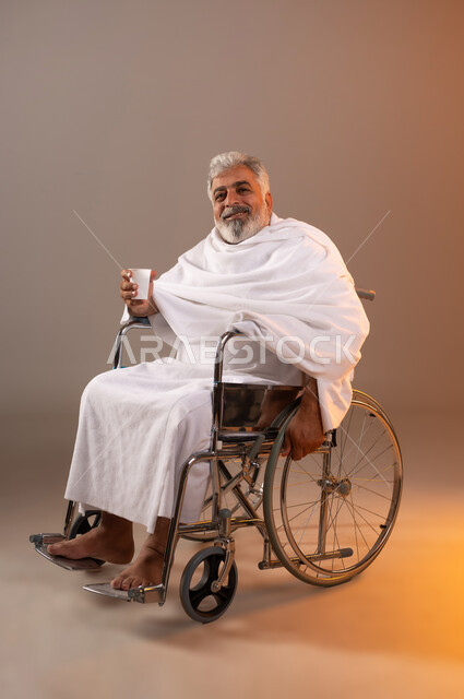 الاستعداد لاداء مناسك الحج والعمره، التقرب الى الله بالاعمال الصالحه، بورتريه لرجل عربي خليجي سعودي كبير بالسن يرتدي ثوب الاحرام يجلس على كرسي متحرك من ذوي الاحتياجات الخاصه يقوم بشرب ماء زمزم، الركن الخامس من أركان الاسلام لمن استطاع اليه سبيلا، خلفيه برتقاليه