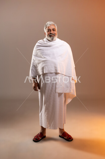 Preparing to perform the rituals of Hajj and Umrah, Islamic matters and worshipers, a portrait of an old man wearing the Ihram dress, standing smiling in front of the camera with gestures of joy and happiness, the fifth pillar of Islam for those who can find a way to it, orange background