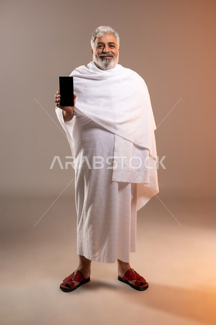الرد على الاتصالات عبر الهاتف المحمول، تعابير السرور بالتواصل مع الاهل والأصدقاء، بورتريه لرجل عربي خليجي سعودي كبير بالسن مبتسم يرتدي ثياب الاحرام يحمل بيده جهاز الهاتف المحمول، التقرب الى الله بأداء فريضة الحج والعمرة واتمام المناسك، خلفيه برتقاليه