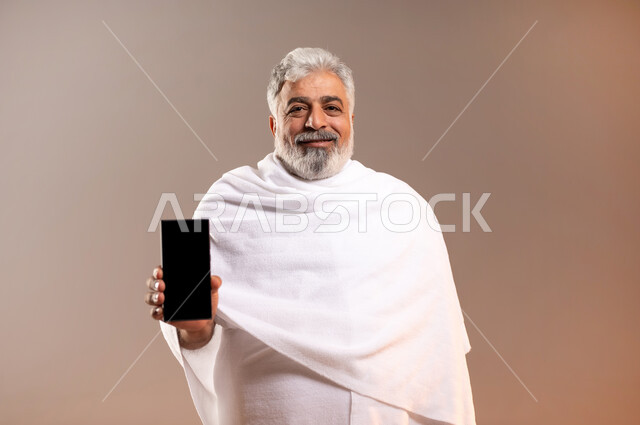 الرد على الاتصالات عبر الهاتف المحمول، تعابير السرور بالتواصل مع الاهل والأصدقاء، بورتريه لرجل عربي خليجي سعودي كبير بالسن مبتسم يرتدي ثياب الاحرام يحمل بيده جهاز الهاتف المحمول، التقرب الى الله بأداء فريضة الحج والعمرة واتمام المناسك، خلفيه برتقاليه