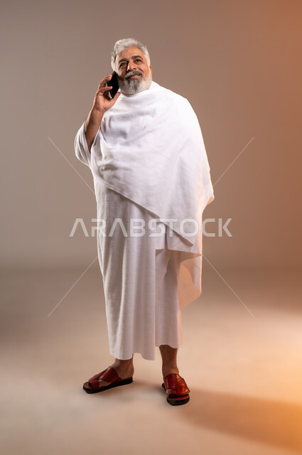 الرد على الاتصالات عبر الهاتف المحمول، تعابير السرور بالتواصل مع الاهل والأصدقاء، بورتريه لرجل عربي خليجي سعودي كبير بالسن مبتسم يرتدي ثياب الاحرام يحمل بيده جهاز الهاتف المحمول، التقرب الى الله بأداء فريضة الحج والعمرة واتمام المناسك، خلفيه برتقاليه