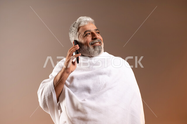 Answering calls via mobile phone, expressions of pleasure in communicating with family and friends, a portrait of an elderly Saudi Gulf Arab man, smiling, wearing Ihram clothes, holding a mobile phone in his hand, getting closer to God by performing the Hajj and Umrah obligations and completing the rituals, orange background.
