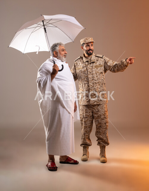 Helping pilgrims and Umrah pilgrims coming to Mecca to perform religious rituals, the gallantry of the security personnel serving the guests of God, a portrait of a Saudi Gulf Arab man wearing a military uniform directing an elderly man wearing Ihram clothes, holding in his hand a white umbrella to protect from the sun’s rays, to reach a place quickly and easily. Orange background