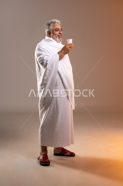 Preparing to perform the rituals of Hajj and Umrah, getting closer to God through good deeds, a portrait of an old Saudi Gulf Arab man wearing the Ihram dress, standing and holding a cup in his hand preparing to drink Zamzam water after performing the rituals of Hajj and Umrah in Mecca, the fifth pillar of Islam for those who have the means to it, background Orange