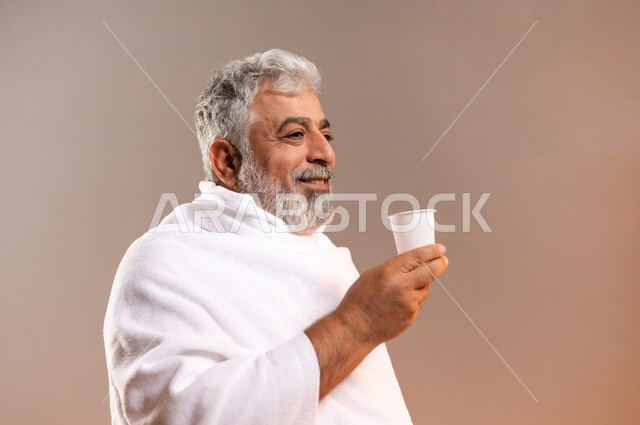 الاستعداد لاداء مناسك الحج والعمره، التقرب الى الله بالاعمال الصالحه، بورتريه لرجل عربي خليجي سعودي كبير بالسن يرتدي ثوب الاحرام يقف ويمسك بيده كوبا استعدادا لشرب ماء زمزم بعد اداء مناسك الحج والعمره في مكه المكرمه، الركن الخامس من أركان الاسلام لمن استطاع اليه سبيلا، خلفيه برتقاليه