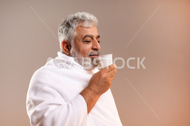 الاستعداد لاداء مناسك الحج والعمره، التقرب الى الله بالاعمال الصالحه، بورتريه لرجل عربي خليجي سعودي كبير بالسن يرتدي ثوب الاحرام يقف ويمسك بيده كوبا استعدادا لشرب ماء زمزم بعد اداء مناسك الحج والعمره في مكه المكرمه، الركن الخامس من أركان الاسلام لمن استطاع اليه سبيلا، خلفيه برتقاليه