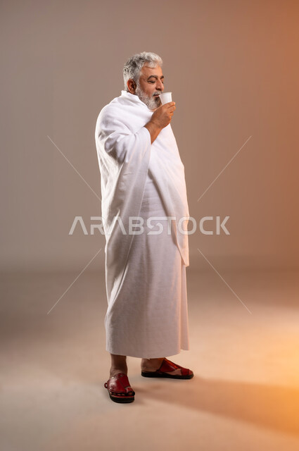 الاستعداد لاداء مناسك الحج والعمره، التقرب الى الله بالاعمال الصالحه، بورتريه لرجل عربي خليجي سعودي كبير بالسن يرتدي ثوب الاحرام يقف ويمسك بيده كوبا استعدادا لشرب ماء زمزم بعد اداء مناسك الحج والعمره في مكه المكرمه، الركن الخامس من أركان الاسلام لمن استطاع اليه سبيلا، خلفيه برتقاليه