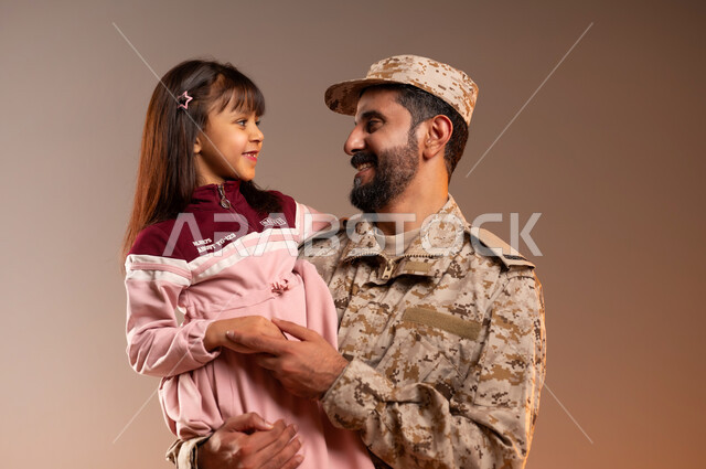 مشاركة اجواء رائعة لعسكري مع صبيه ، شهامه رجال الامن في التعامل مع الاطفال، بورتريه لرجل عربي خليجي سعودي يرتدي الزي العسكري يحمل فتاه صغيره ترتدي فستانا باللون الزهري ويضمها اليه بأيماءات الطمأنينه والحنان، جيش الدفاع الوطني السعودي، خلفيه برتقاليه