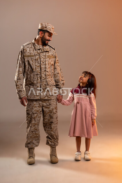 مشاركة اجواء رائعة لعسكري مع صبيه ، شهامه رجال الامن في التعامل مع الاطفال، بورتريه لرجل عربي خليجي سعودي يرتدي الزي العسكري يقف بجانب فتاه صغيره ترتدي فستانا باللون الزهري يمسك بيدها بأيماءات التحيه والطمأنينه، جيش الدفاع الوطني السعودي، خلفيه برتقاليه