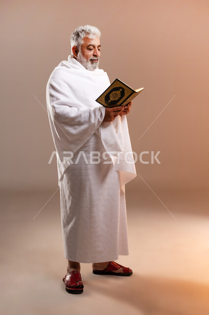 تدبر وتمعن آيات كتاب الله, التقرب الى الله بقراءة القرآن, بورتريه لرجل عربي سعودي خليجي كبير السن يرتدي لباس الاحرام يحمل بيده كتاب الله (القرآن الكريم) ، اداء مناسك الحج والعمرة ، طاعات وعبادات، اسلاميات وعباده، خلفيه برتقاليه