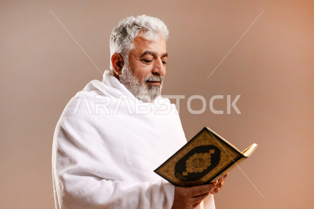 تدبر وتمعن آيات كتاب الله, التقرب الى الله بقراءة القرآن, بورتريه لرجل عربي سعودي خليجي كبير السن يرتدي لباس الاحرام يحمل بيده كتاب الله (القرآن الكريم) ، اداء مناسك الحج والعمرة ، طاعات وعبادات، اسلاميات وعباده، خلفيه برتقاليه