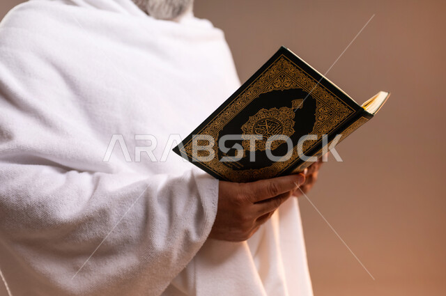 تدبر وتمعن آيات كتاب الله, التقرب الى الله بقراءة القرآن, بورتريه لرجل عربي سعودي خليجي كبير السن يرتدي لباس الاحرام يحمل بيده كتاب الله (القرآن الكريم) ، اداء مناسك الحج والعمرة ، طاعات وعبادات، اسلاميات وعباده، خلفيه برتقاليه