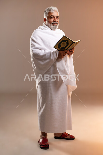 تدبر وتمعن آيات كتاب الله, التقرب الى الله بقراءة القرآن, بورتريه لرجل عربي سعودي خليجي كبير السن يرتدي لباس الاحرام يحمل بيده كتاب الله (القرآن الكريم) ، اداء مناسك الحج والعمرة ، طاعات وعبادات، اسلاميات وعباده، خلفيه برتقاليه
