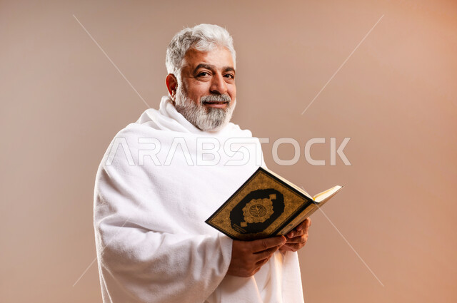 تدبر وتمعن آيات كتاب الله, التقرب الى الله بقراءة القرآن, بورتريه لرجل عربي سعودي خليجي كبير السن يرتدي لباس الاحرام يحمل بيده كتاب الله (القرآن الكريم) ، اداء مناسك الحج والعمرة ، طاعات وعبادات، اسلاميات وعباده، خلفيه برتقاليه