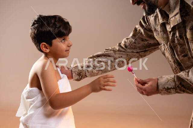 مشاركة اجواء رائعة لعسكري مع صبي ، تقديم الحلوى للاطفال وادخال البهجه والسرور الى قلوبهم، بورتريه لرجل عربي خليجي سعودي يرتدي الزي العسكري يقوم بتقديم حلوى المصاص لصبي يرتدي ثوب الاحرام، تأديه مناسك الحج والعمره بيسر وطمأنينه، خلفيه برتقاليه