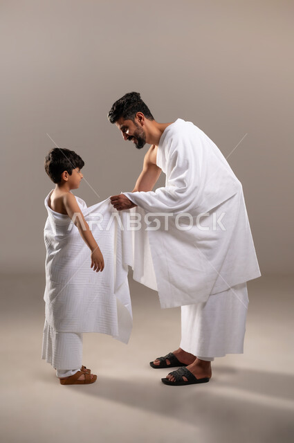 الاستعداد لاداء مناسك الحج والعمره والتقرب الى الله بالاعمال الصالحه، بورتريه لرجل عربي خليجي سعودي يرتدي ثوب الاحرام يقوم بتعليم ابنه الصبي كيفيه ارتداء زي الاحرام، تأديه الركن الخامس من أركان الاسلام لمن استطاع اليه سبيلا، اسلاميات وعباده، خلفيه رماديه