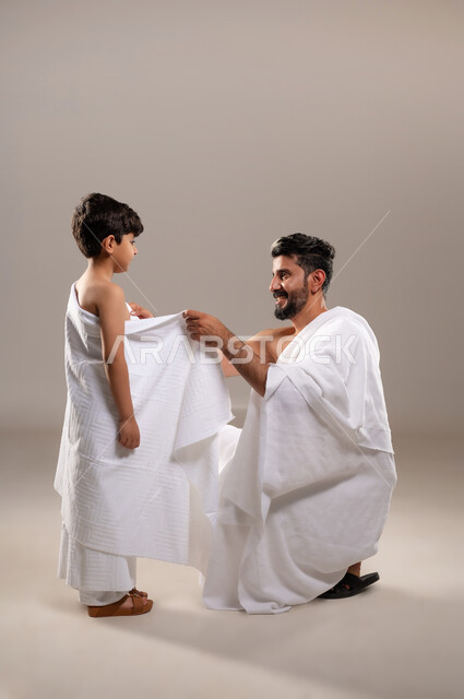 الاستعداد لاداء مناسك الحج والعمره والتقرب الى الله بالاعمال الصالحه، بورتريه لرجل عربي خليجي سعودي يرتدي ثوب الاحرام يقوم بتعليم ابنه الصبي كيفيه ارتداء زي الاحرام، تأديه الركن الخامس من أركان الاسلام لمن استطاع اليه سبيلا، اسلاميات وعباده، خلفيه رماديه