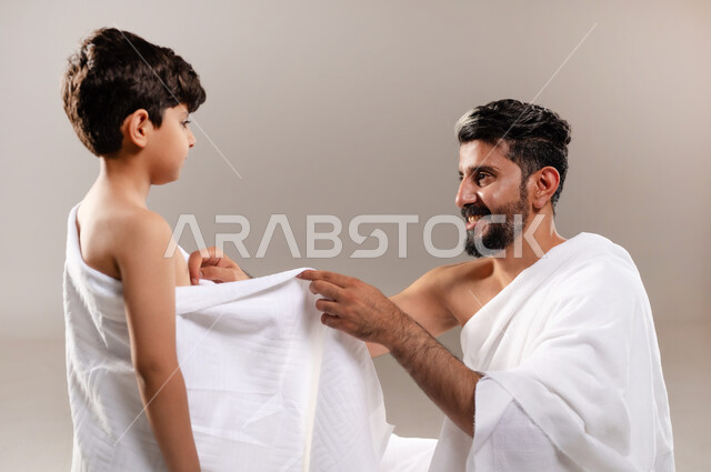 الاستعداد لاداء مناسك الحج والعمره والتقرب الى الله بالاعمال الصالحه، بورتريه لرجل عربي خليجي سعودي يرتدي ثوب الاحرام يقوم بتعليم ابنه الصبي كيفيه ارتداء زي الاحرام، تأديه الركن الخامس من أركان الاسلام لمن استطاع اليه سبيلا، اسلاميات وعباده، خلفيه رماديه
